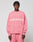 A person with short, twisted hair wears the LIVE FAST DIE YOUNG Live Fast Boucle Sweater in an oversize pink fit with “LIVE FAST” in bold white letters, paired with matching pants against a plain, light background.