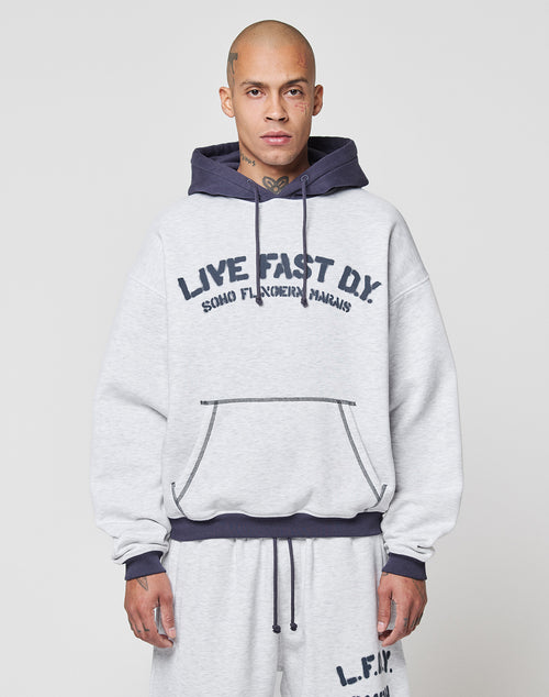 A person with a shaved head wears the LFDY District Hooded by LIVE FAST DIE YOUNG, with dark blue contrast drawstrings, a kangaroo pocket, and logo print. They pair it with matching gray sweatpants.