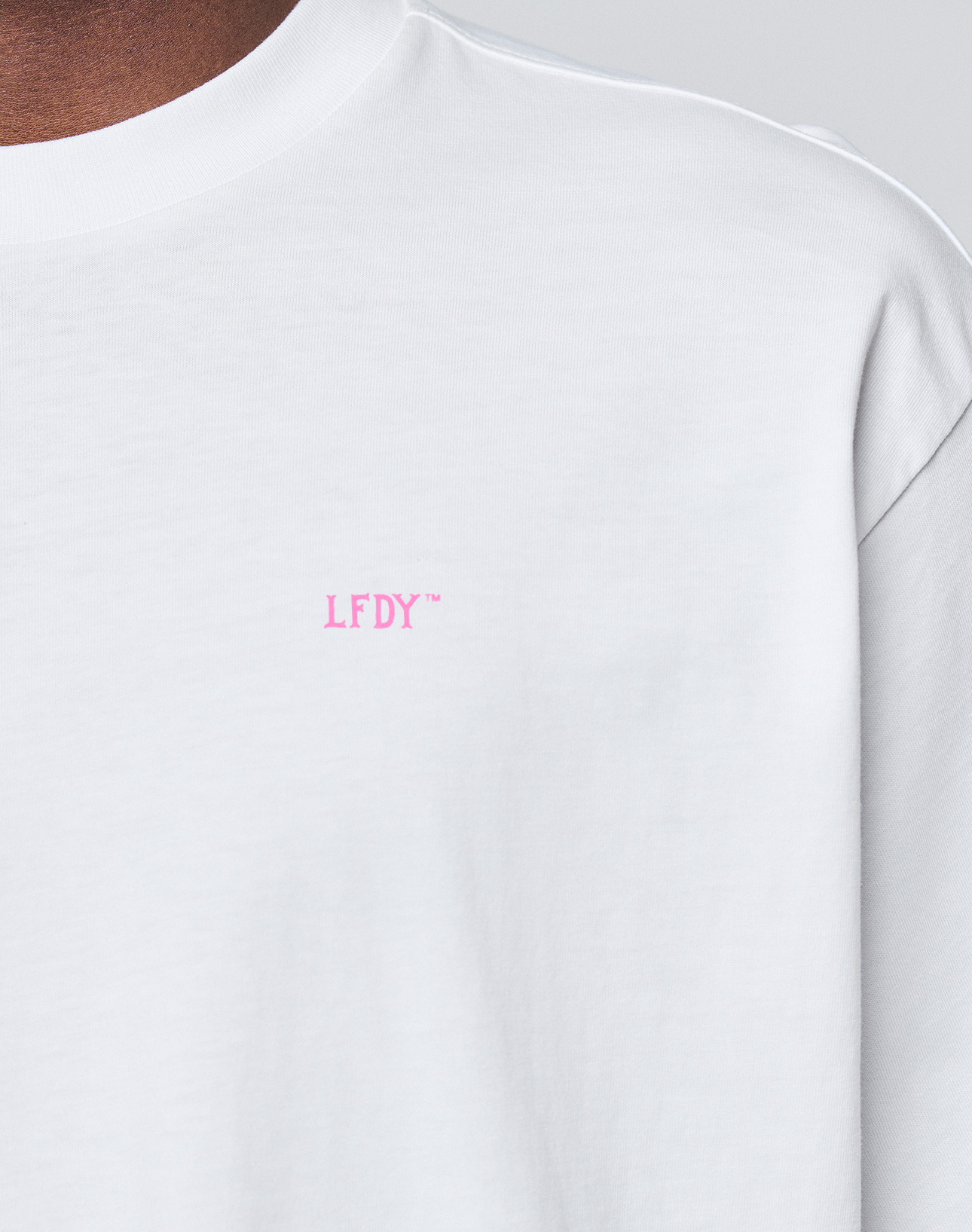 Close-up of the Basic Evidence Tee by LIVE FAST DIE YOUNG: a white, 100% Baumwolle T-shirt with small pink LFDY™ text on the upper left chest. Oversize fit. Neutral background shows part of neck and shoulder.