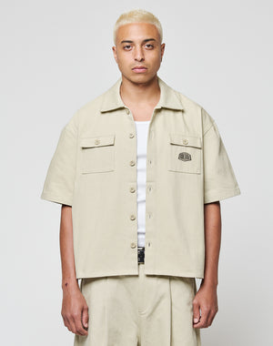 A person with short platinum blond hair stands against a plain background, wearing the LFDY Twill Shirt by LIVE FAST DIE YOUNG over a white top and matching beige pants, looking directly at the camera with a neutral expression.