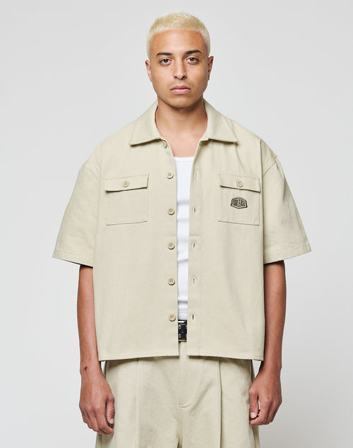A person with short platinum blond hair stands against a plain background, wearing the LFDY Twill Shirt by LIVE FAST DIE YOUNG over a white top and matching beige pants, looking directly at the camera with a neutral expression.