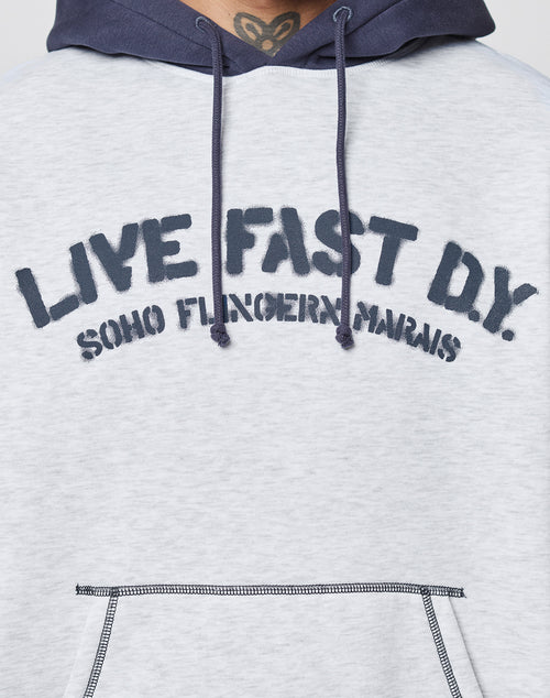 Close-up of a person wearing a light gray LFDY District Hooded by LIVE FAST DIE YOUNG, featuring dark blue drawstrings and hood. "LIVE FAST D.Y. SOHO FUNGERIN MARAIS" is boldly printed on the front.