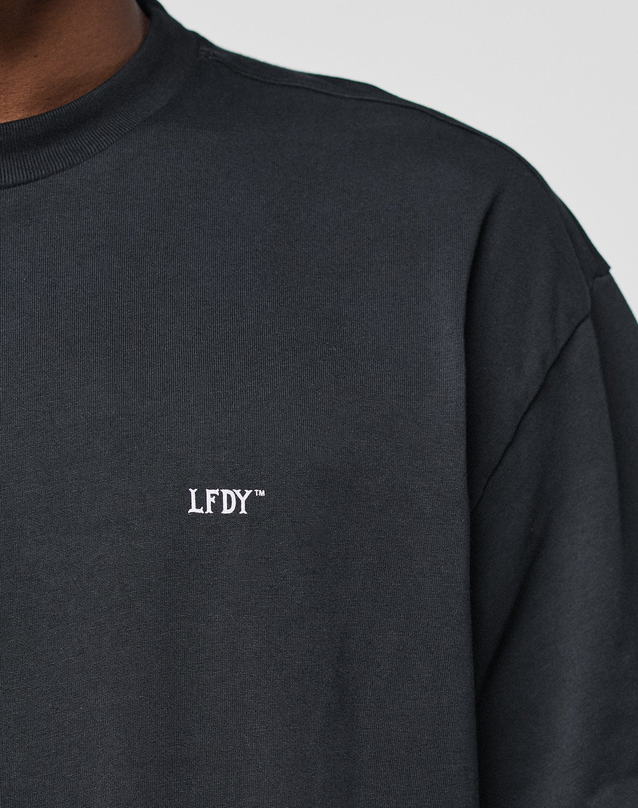 Close-up of a person wearing the LIVE FAST DIE YOUNG Basic Trademark Tee, featuring an oversized fit and small white LFDY™ embroidery on the chest. Only the upper torso and part of the neck are visible. Made from 100% cotton.