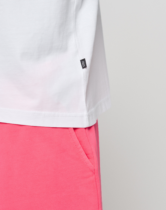 Close-up of a person wearing the LIVE FAST DIE YOUNG Basic Evidence Tee in white Oversize Fit (100% Baumwolle) with a black tag on the side seam and bright pink pants, hand in pocket, against a plain light background.