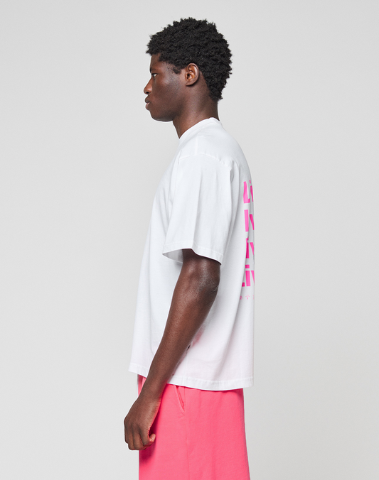 A person stands in profile against a plain background, wearing a LIVE FAST DIE YOUNG Basic Evidence Tee—an oversize fit white T-shirt with pink front and back print—and bright pink shorts.