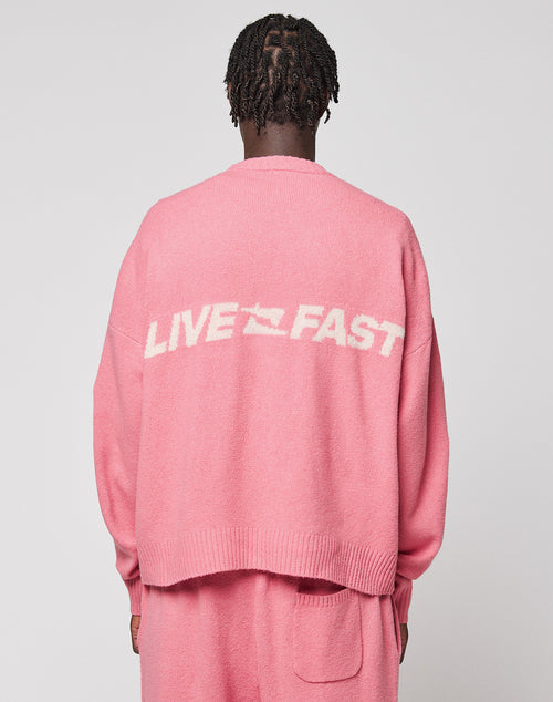 Seen from behind, a person with short braided hair wears the LIVE FAST DIE YOUNG "Live Fast Boucle Sweater," an oversized pink knit with "LIVE FAST" in white on the back, paired with matching pink pants against a plain light background.