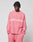 A person wears the LIVE FAST DIE YOUNG Live Fast Boucle Sweater in oversize-fit pink with “LIVE FAST” in white, paired with matching pink pants, standing hands in pockets and looking down against a plain light background.