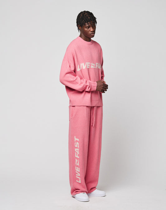 A person wears the Live Fast Boucle Sweater by LIVE FAST DIE YOUNG and matching pink wide-leg pants, both featuring "LIVE FAST" in white, styled with white sneakers against a plain light background.