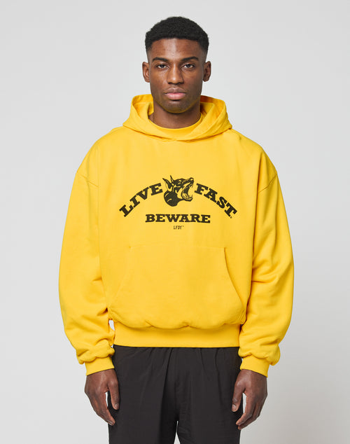 A man wearing a bright yellow LIVE FAST DIE YOUNG Live Fast Beware Hooded with LIVE FAST BEWARE and a fierce animal graphic on the front stands against a plain light gray background.