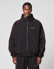 A man faces forward wearing the LIVE FAST DIE YOUNG Live Fast Trackjacket and matching pants with orange piping, reflektierender Druck, and LIVE FAST printed on the chest and leg, set against a plain light gray background. | model
