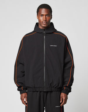 A man faces forward wearing the LIVE FAST DIE YOUNG Live Fast Trackjacket and matching pants with orange piping, reflektierender Druck, and LIVE FAST printed on the chest and leg, set against a plain light gray background.