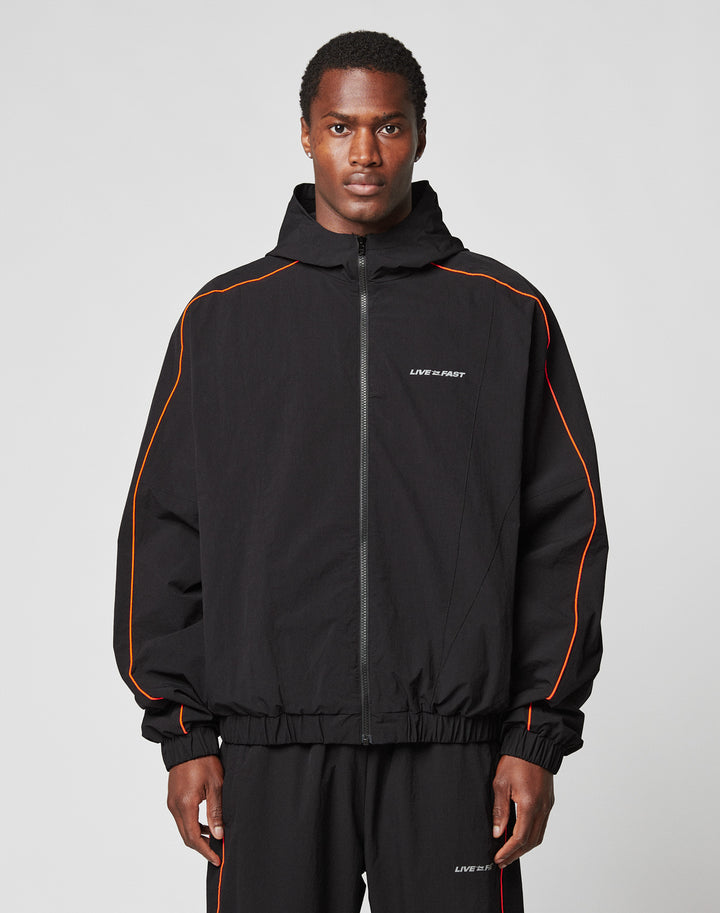A man faces forward wearing the LIVE FAST DIE YOUNG Live Fast Trackjacket and matching pants with orange piping, reflektierender Druck, and LIVE FAST printed on the chest and leg, set against a plain light gray background. | model