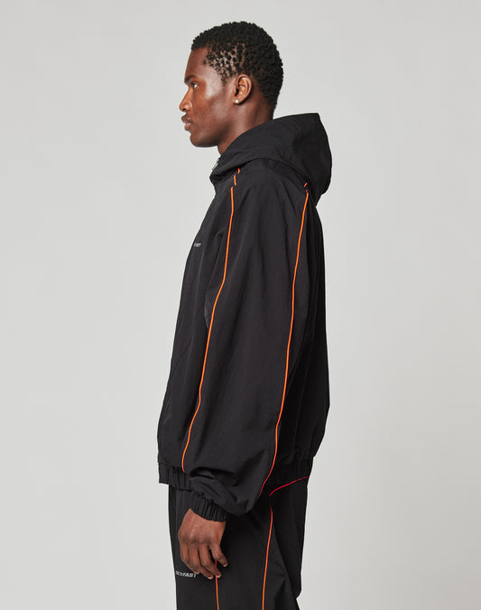 A man stands in profile wearing the LIVE FAST DIE YOUNG Live Fast Trackjacket, a black hooded nylon jacket with orange piping and elastic cuffs, paired with matching black pants against a plain light background.