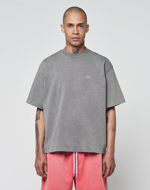A person with a shaved head and tattoos wears the LIVE FAST DIE YOUNG Basic Trademark Tee in gray and pink shorts, standing against a light gray background, facing forward with a neutral expression.