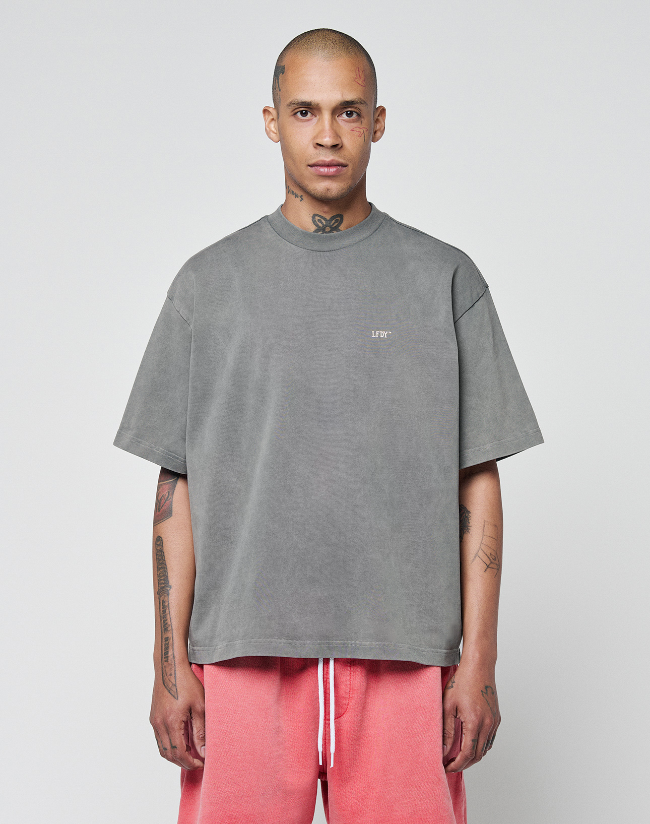 A person with a shaved head and visible tattoos wears the LIVE FAST DIE YOUNG Basic Trademark Tee in oversize fit and gray, paired with pink drawstring shorts, standing against a plain white background and looking straight ahead.