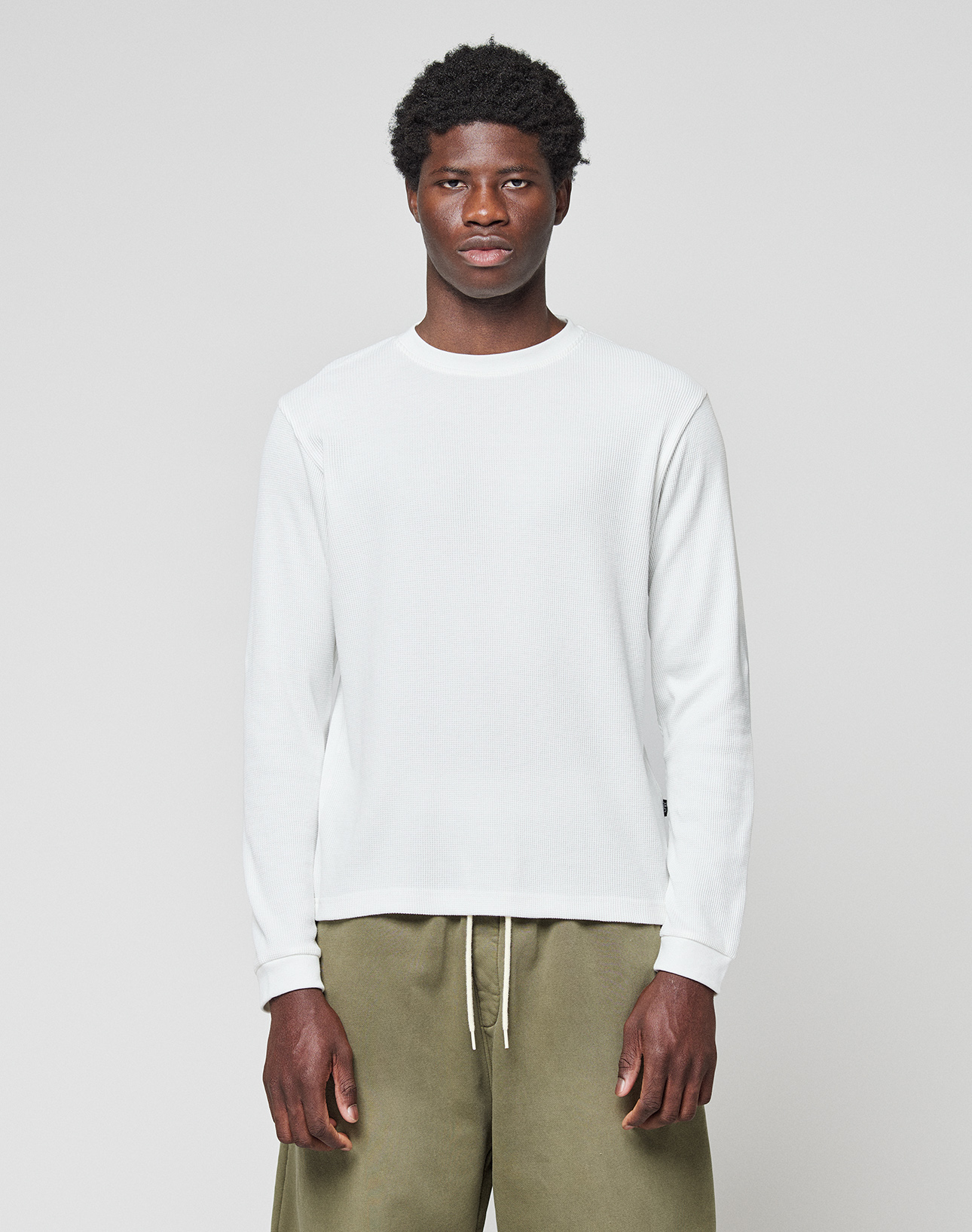 A man faces forward, arms relaxed, wearing the LIVE FAST DIE YOUNG Basic Waffle Longsleeve in white and olive green pants, against a plain background. The shirt features an oversize fit and flag-label detail.