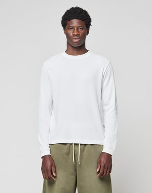 A man faces forward, arms relaxed, wearing the LIVE FAST DIE YOUNG Basic Waffle Longsleeve in white and olive green pants, against a plain background. The shirt features an oversize fit and flag-label detail.