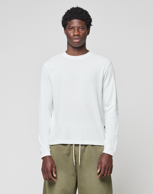 A man faces forward, arms relaxed, wearing the LIVE FAST DIE YOUNG Basic Waffle Longsleeve in white and olive green pants, against a plain background. The shirt features an oversize fit and flag-label detail.