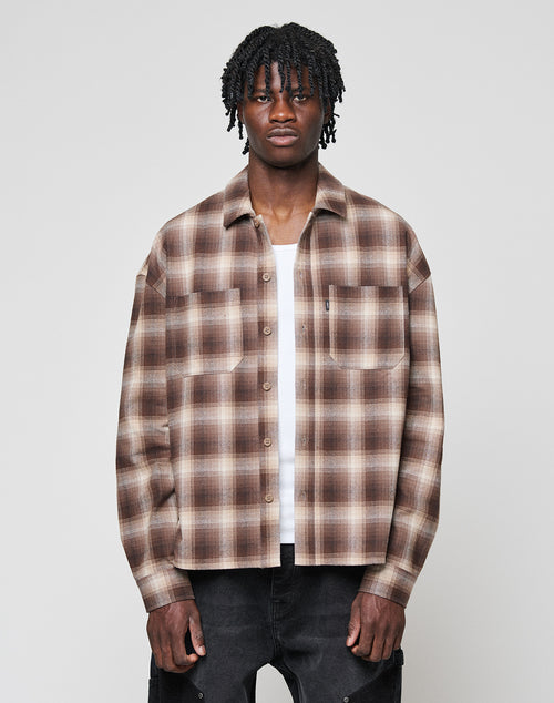 A man with short dreadlocks wears a LIVE FAST DIE YOUNG Vintage Flannel in brown and beige plaid over a white T-shirt and black pants, standing against a plain light gray background.