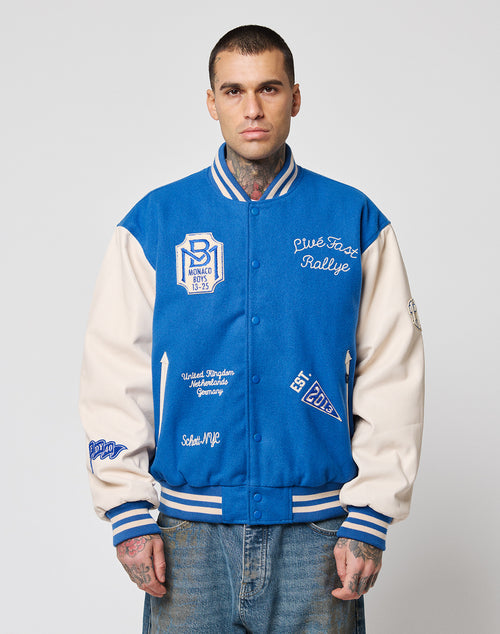 A man with short hair and tattoos wears the LIVE FAST DIE YOUNG LFDY x Schott NYC Varsity Jacket with embroidered patches, cowhide leather sleeves, and Monaco Rallye text over light blue jeans, standing against a plain white background.