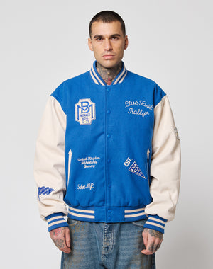 A man with short hair and tattoos wears the LIVE FAST DIE YOUNG LFDY x Schott NYC Varsity Jacket with embroidered patches, cowhide leather sleeves, and Monaco Rallye text over light blue jeans, standing against a plain white background.