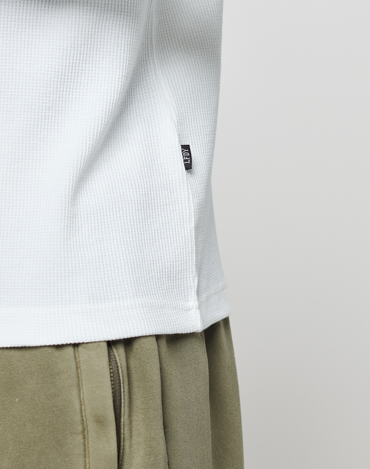 A close-up shows a person wearing the LIVE FAST DIE YOUNG Basic Waffle Longsleeve in an oversize white textured Baumwolle, featuring a small black flag-label on the side seam, paired with light olive-green pants against a plain background.