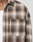 A close-up shows someone wearing the LIVE FAST DIE YOUNG Vintage Flannel, an oversize fit brown and beige plaid shirt in 100% Baumwolle, layered over a white top. The shirt has a front pocket with a black tag; image cropped at neck and mid-chest.