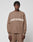 A person with short, braided hair wears the LIVE FAST DIE YOUNG Live Fast Boucle Sweater—an oversize brown piece with "LIVE FAST" in white across the chest—paired with matching brown pants against a plain light background.