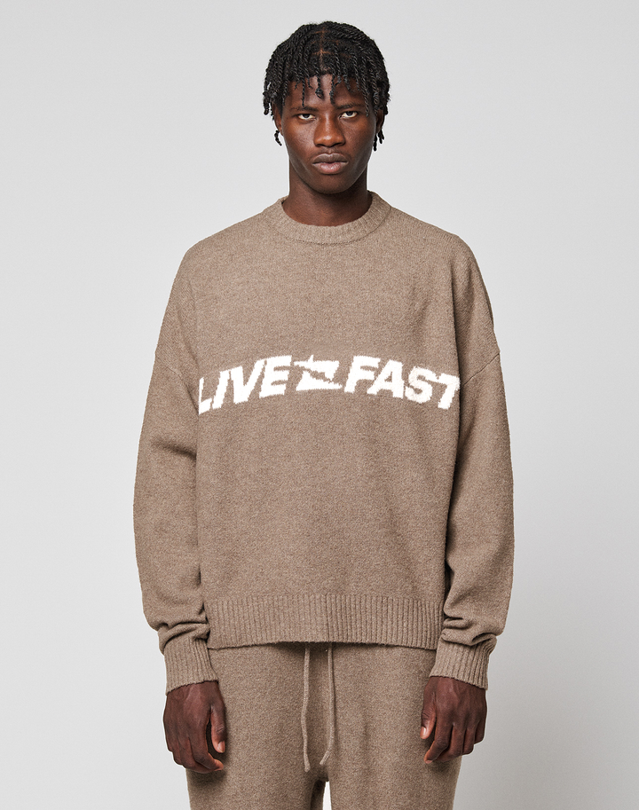 A person wears the LIVE FAST DIE YOUNG Live Fast Boucle Sweater, an oversized beige knit with "LIVE FAST" in bold white letters, paired with matching pants against a plain background.