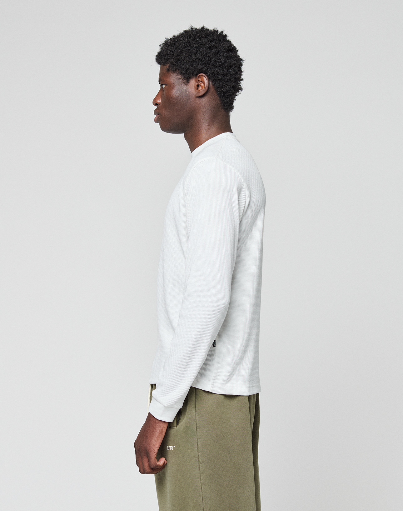 A man with short curly hair stands in profile, wearing the LIVE FAST DIE YOUNG Basic Waffle Longsleeve in oversize fit and olive green pants, set against a plain light grey background.