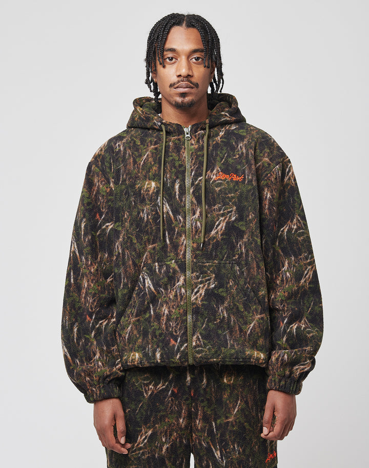 A person with braided hair wears the LIVE FAST DIE YOUNG LFDY Fleece Hooded Zip in a camouflage pattern with kapuze mit kordeln and red embroidered text on the left chest, paired with matching pants against a plain white background. | model