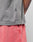 A person wearing a gray Basic Trademark Tee by LIVE FAST DIE YOUNG with a small black tag and pink shorts stands with one hand in their pocket, revealing part of their colorful arm tattoo.