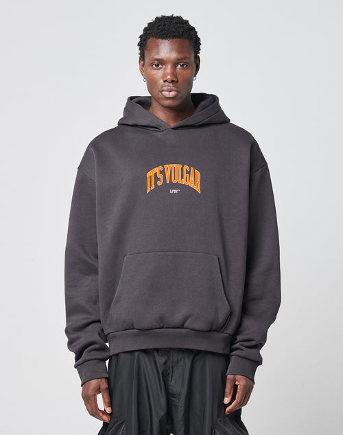 A person stands facing forward wearing a loose-fitting LIVE FAST DIE YOUNG Its Vulgar Center Hooded sweatshirt in an Oversize Fit, with ITS VULGAR printed in orange and yellow on the front against a plain light gray background.