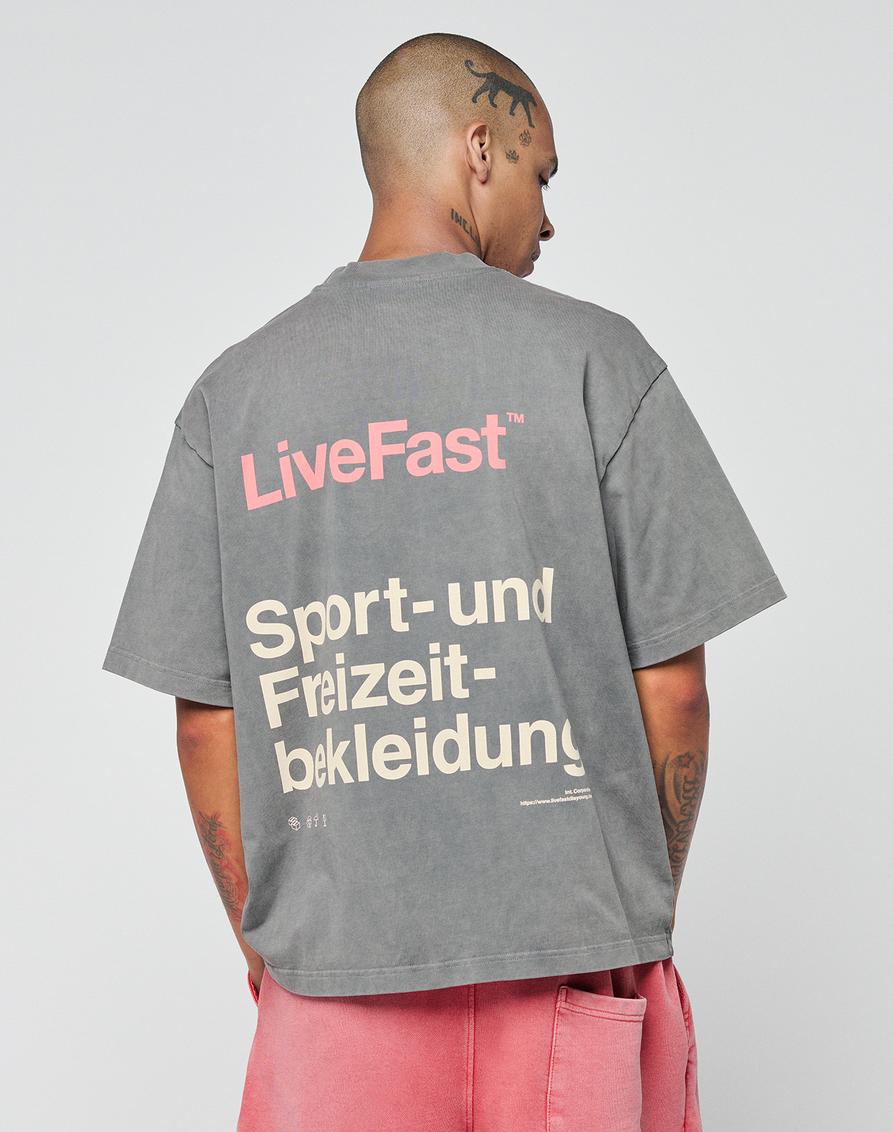 A person with a shaved head, visible tattoos, and hands behind their back wears the LIVE FAST DIE YOUNG Basic Trademark Tee in gray and pink shorts—a unique look against a simple backdrop.