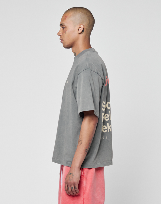 A person with a shaved head and facial tattoos stands in profile, wearing the Basic Trademark Tee by LIVE FAST DIE YOUNG in gray oversize fit with back text and pink shorts, their unique look contrasting the plain white background.