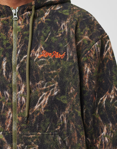Close-up of a person wearing the LFDY Fleece Hooded Zip by LIVE FAST DIE YOUNG, a camouflage fleece jacket with drawstring hood, zipper, oversize fit, and an orange embroidered Live Fast logo on the left chest.