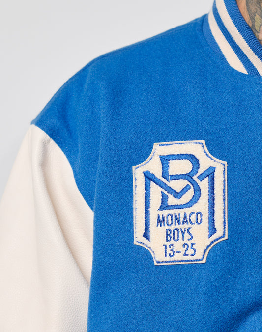Close-up of the LIVE FAST DIE YOUNG x Schott NYC Varsity Jacket in blue and white, featuring cowhide leather sleeves and a chenille Monaco Boys 13-25 patch with a stylized MB logo on the chest.