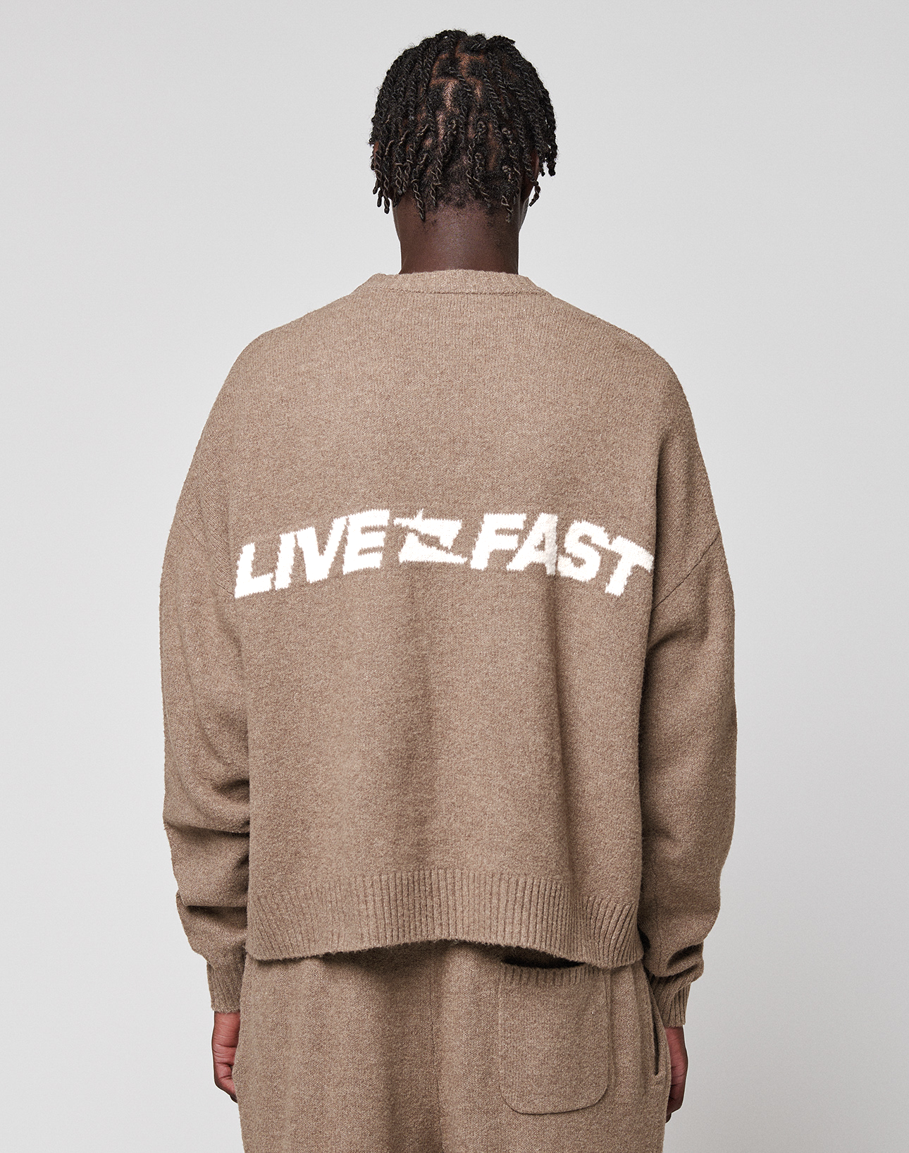 A person with short, twisted hair is seen from behind wearing the LIVE FAST DIE YOUNG Live Fast Boucle Sweater in light brown with "LIVE FAST" in white, paired with matching knit pants. The background is plain gray.