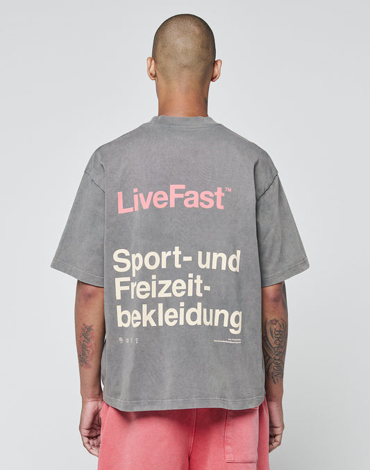 A person with a shaved head (seen from behind) wears an oversize washed gray LIVE FAST DIE YOUNG Basic Trademark Tee with “LiveFast” in pink and “Sport- und Freizeitbekleidung” in beige on the back, styled with pink shorts.