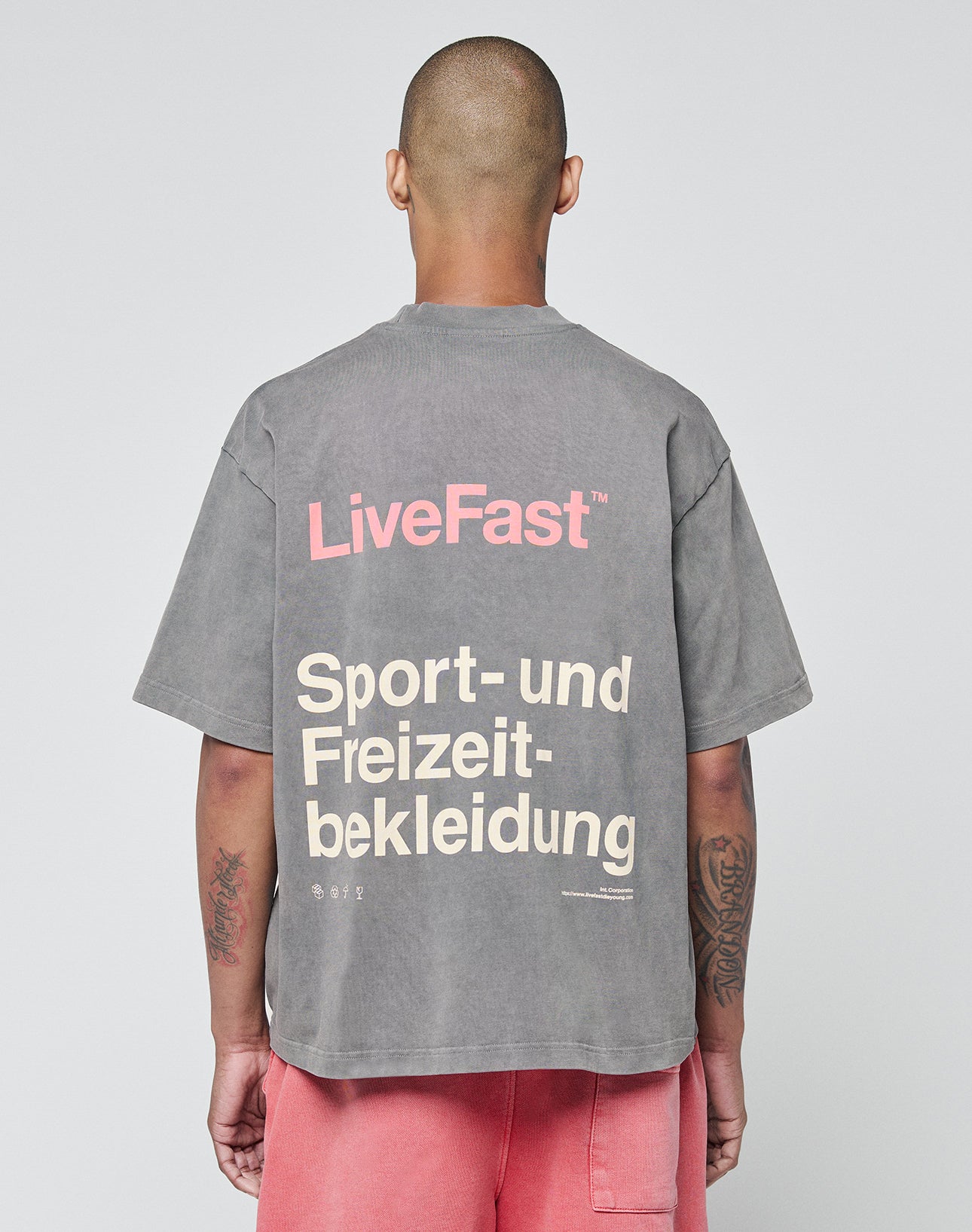 A person with a shaved head (seen from behind) wears an oversize washed gray LIVE FAST DIE YOUNG Basic Trademark Tee with “LiveFast” in pink and “Sport- und Freizeitbekleidung” in beige on the back, styled with pink shorts.