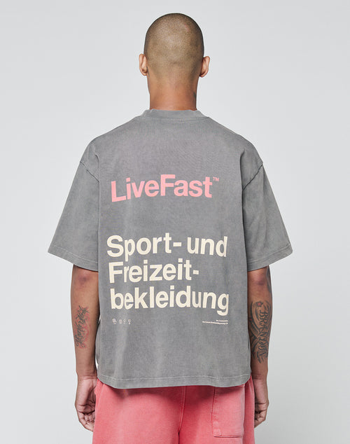 A person with a shaved head (seen from behind) wears an oversize washed gray LIVE FAST DIE YOUNG Basic Trademark Tee with “LiveFast” in pink and “Sport- und Freizeitbekleidung” in beige on the back, styled with pink shorts.
