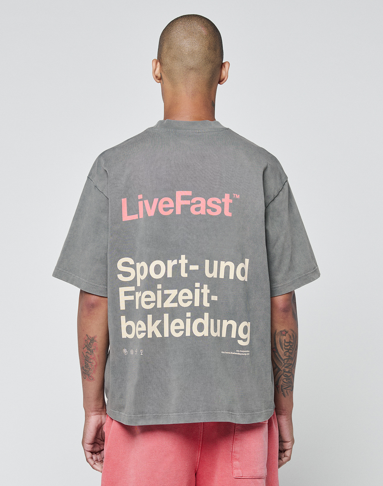Seen from behind, a person with a shaved head wears the LIVE FAST DIE YOUNG Basic Trademark Tee in gray with pink LiveFast and beige Sport- und Freizeitbekleidung text. Oversize fit and pink shorts create a unique look against a plain background.