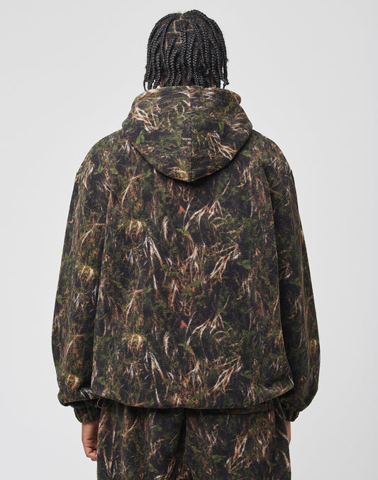 A person with braided hair, seen from behind, wears the LFDY Fleece Hooded Zip by LIVE FAST DIE YOUNG and matching camouflage pants in green and brown hues. The Oversize Fit gives a relaxed vibe against the light background.
