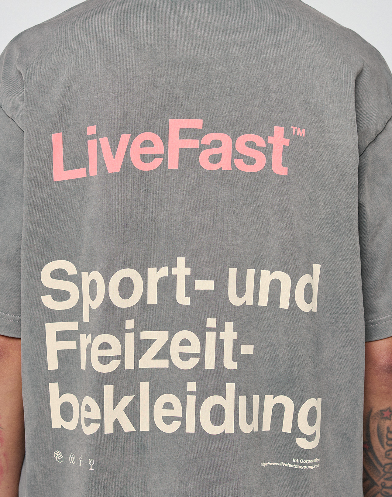 A person seen from the back wears a gray Basic Trademark Tee by LIVE FAST DIE YOUNG, featuring "LiveFast" in pink above "Sport- und Freizeitbekleidung" in beige. Small logos and website text appear near the hem for a unique look.