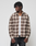 A person stands against a plain background in the LIVE FAST DIE YOUNG Vintage Flannel—an oversize fit brown and beige plaid 100% Baumwolle shirt—layered over a white t-shirt and black pants, looking to the side neutrally.