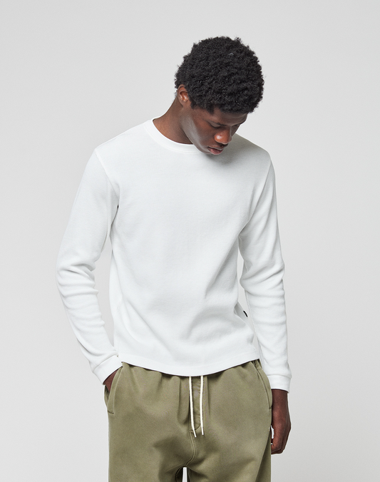 A man in a LIVE FAST DIE YOUNG Basic Waffle Longsleeve (white, oversized fit) and olive green drawstring Baumwolle pants stands with hands in pockets against a plain light gray background, looking down.