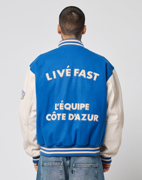 A person with short hair, seen from behind, wears the LIVE FAST DIE YOUNG x Schott NYC Varsity Jacket featuring chenille patches, cowhide leather sleeves, and large white LIVÉ FAST LÉQUIPE CÔTE DAZUR text on the back.