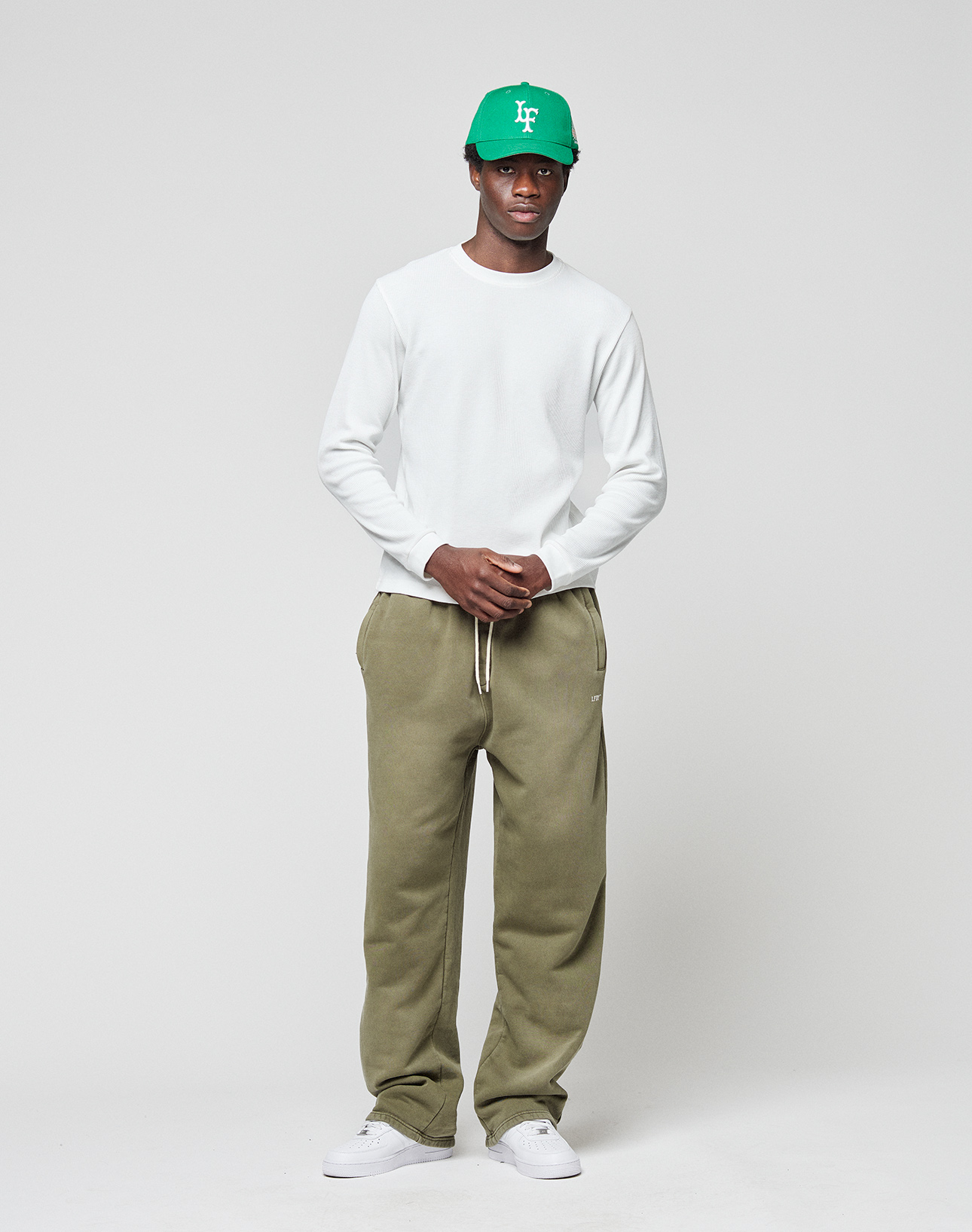 A man stands against a plain background wearing the LIVE FAST DIE YOUNG Basic Waffle Longsleeve in white, paired with olive green sweatpants, white sneakers, and a green baseball cap.