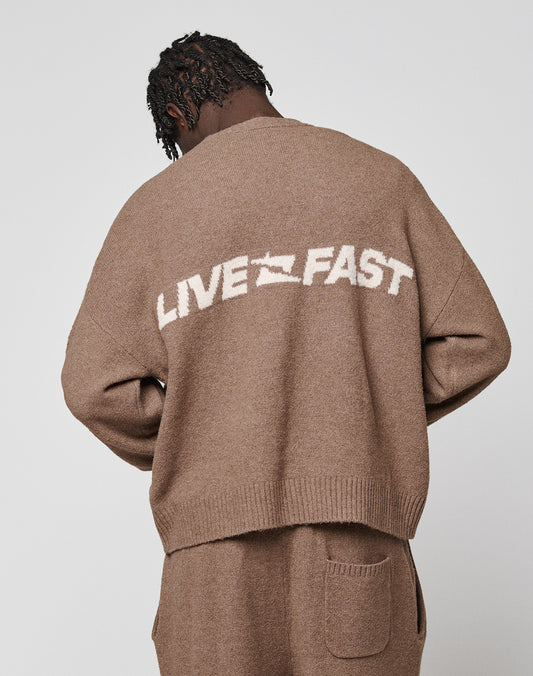 A person with short, twisted hair faces away, wearing the LIVE FAST DIE YOUNG Live Fast Boucle Sweater in an oversized fit with a graphic on the back, paired with matching brown pants.