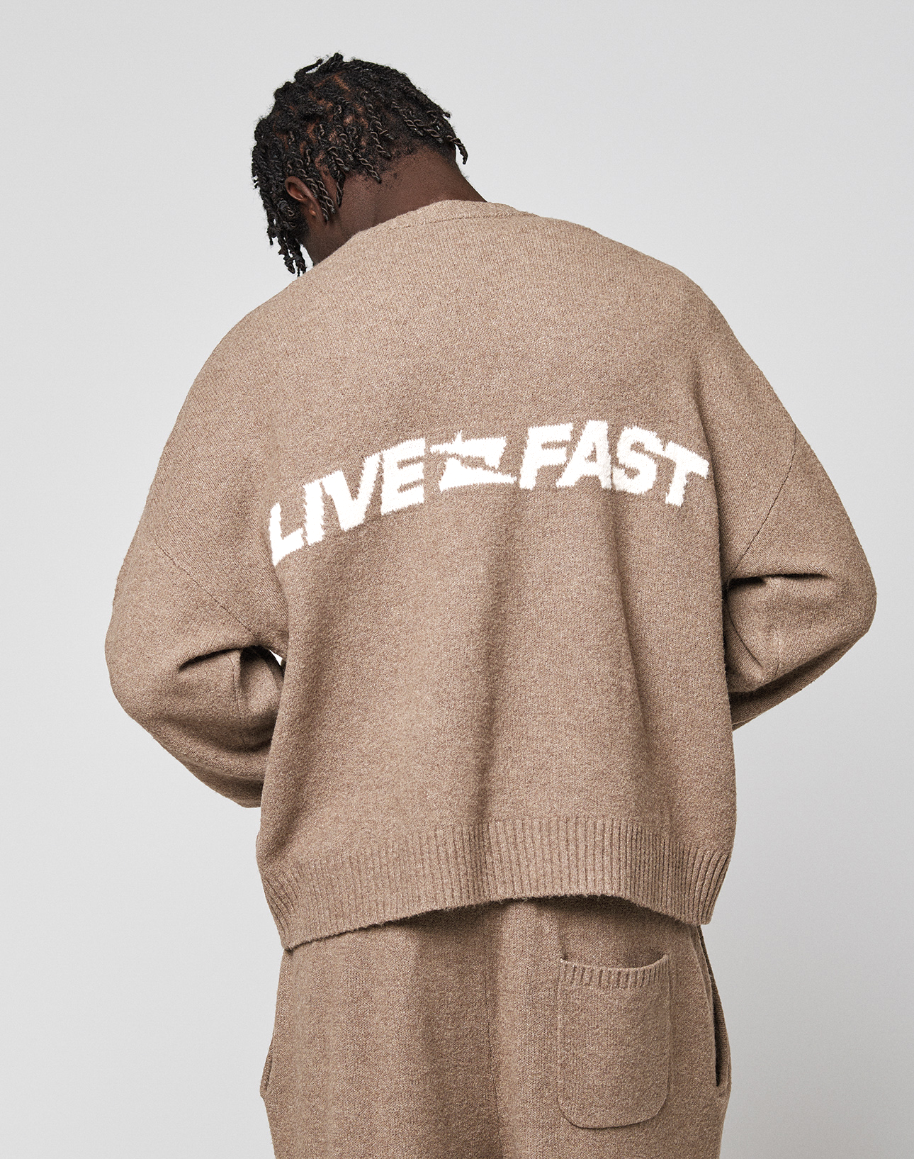 A person with short dreadlocks faces away, wearing the oversized brown LIVE FAST DIE YOUNG "Live Fast Boucle Sweater." The back of the sweater has bold white "LIVE FAST" text and a stylized graphic between the words.